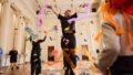 Whirlwind production photo: Laura May Grogan. Two Polyglot artists leap in the air as colourful confetti falls. A child stands in the corner and watches on.