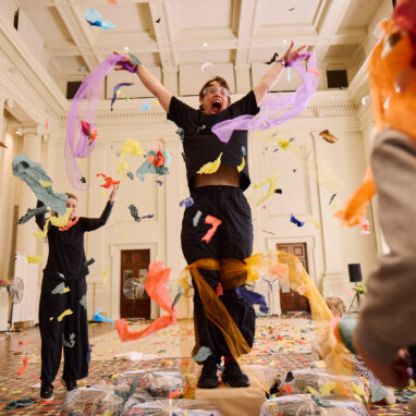 Whirlwind production photo: Laura May Grogan. Two Polyglot artists leap in the air as colourful confetti falls. A child stands in the corner and watches on.