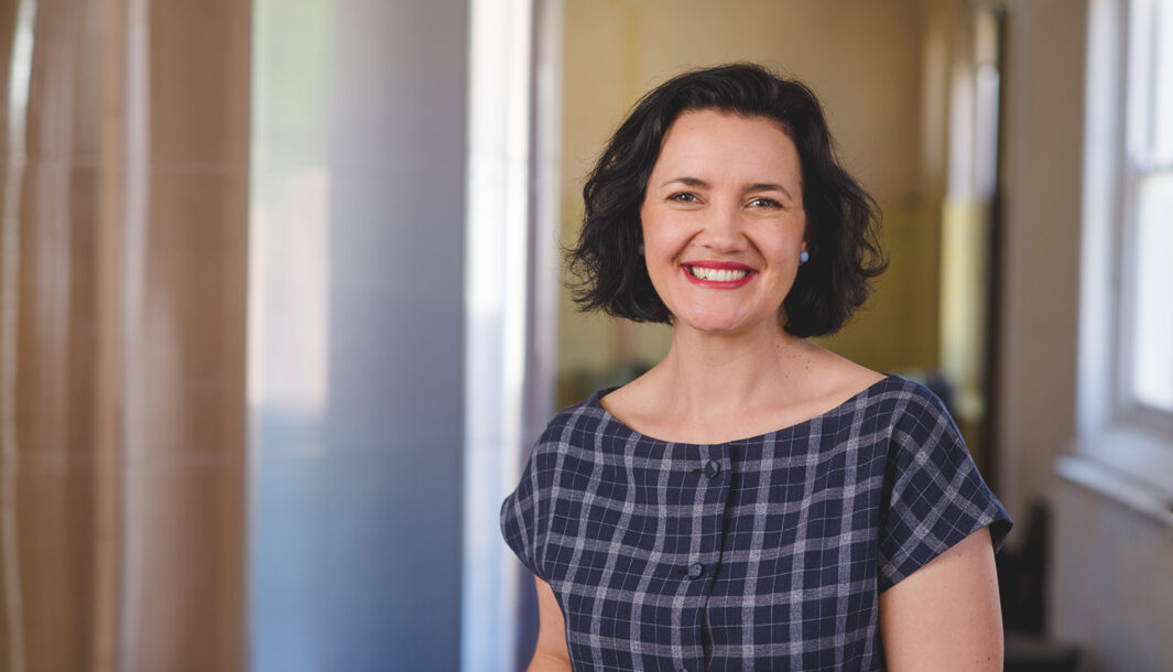 Photo of Kath Fyffe, by Theresa Harrison. Kath smiles at the camera. She has short dark hair, and wears a blue checked dress. She stands in the Polyglot office space.