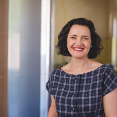 Photo of Kath Fyffe, by Theresa Harrison. Kath smiles at the camera. She has short dark hair, and wears a blue checked dress. She stands in the Polyglot office space.