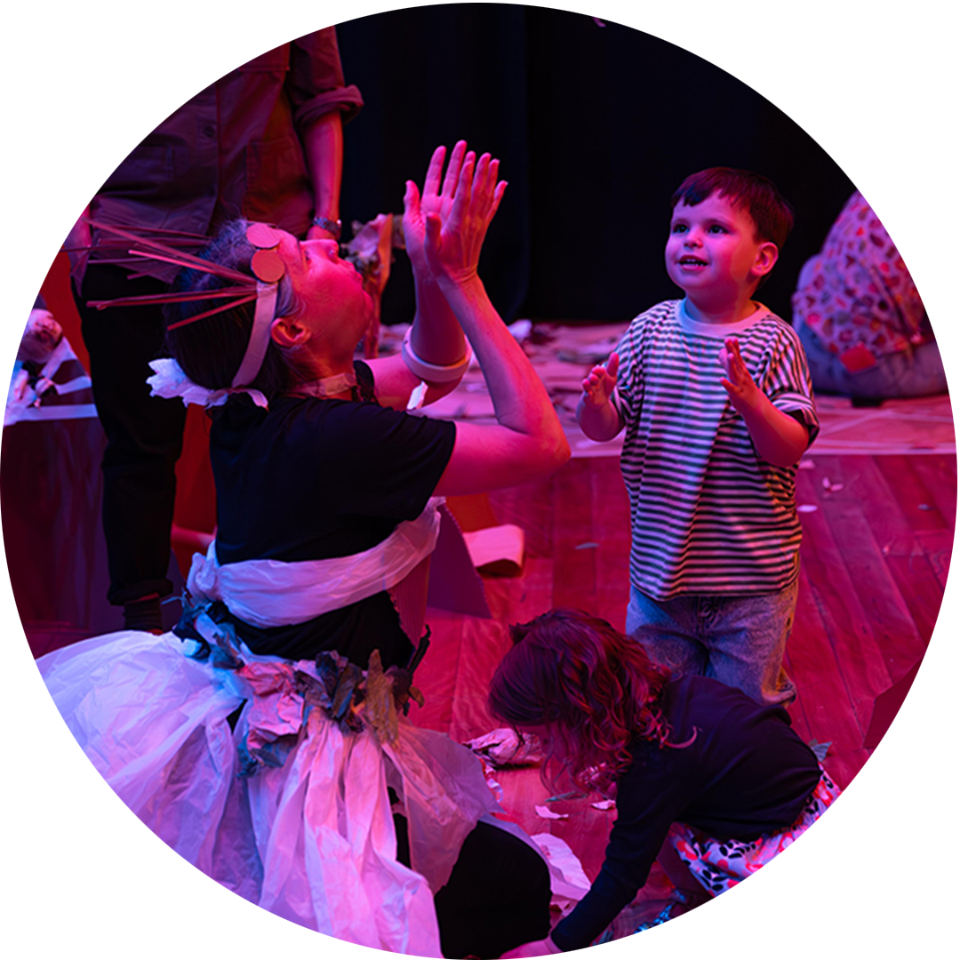 Paper Planet production photo: Katje Ford, Sydney Opera House. A Polyglot artist in an intricate, handmade paper costume engages with a small child, throwing pieces of paper into the air. They are in a darkened, theatrically lit space.