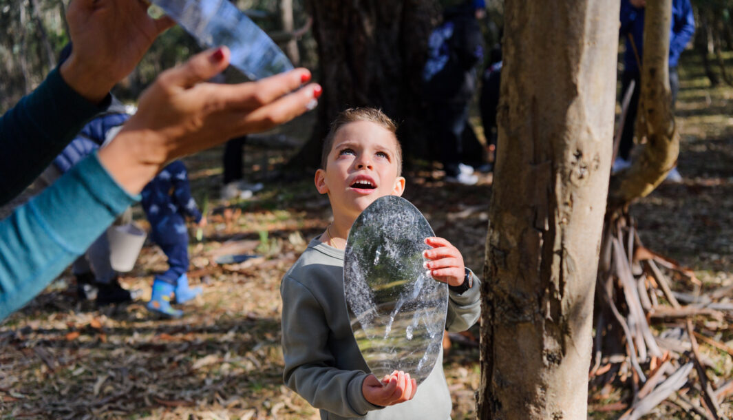 Forest production photo: Sarah Walker. A child stands in a bushland clearing, holding an oblong mirror to reflect light. A Polyglot artist does the same (only their hands and the mirror are visible). Children and families play among the trees in the background.
