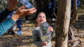 Forest production photo: Sarah Walker. A child stands in a bushland clearing, holding an oblong mirror to reflect light. A Polyglot artist does the same (only their hands and the mirror are visible). Children and families play among the trees in the background.