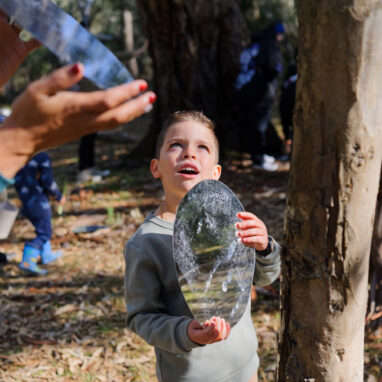 Forest production photo: Sarah Walker. A child stands in a bushland clearing, holding an oblong mirror to reflect light. A Polyglot artist does the same (only their hands and the mirror are visible). Children and families play among the trees in the background.