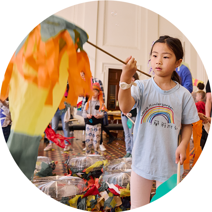 Whirlwind production photo: Laura May Grogan. A child creates and plays with a colourful fish-shaped kite. Children and families create and play in the background, with colourful paper and silver fans.
