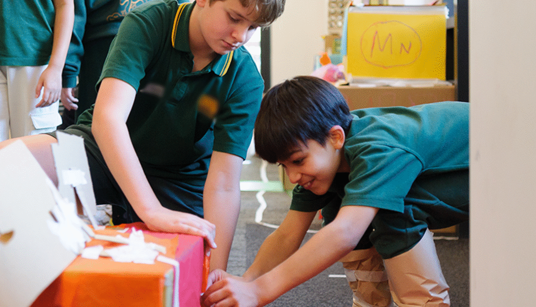 Wish Street production photo: Sarah Walker. Two children work together to attach paper to a box using tape. They both wear green school uniforms.