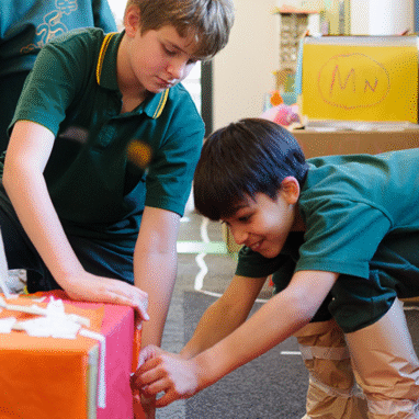 Wish Street production photo: Sarah Walker. Two children work together to attach paper to a box using tape. They both wear green school uniforms.