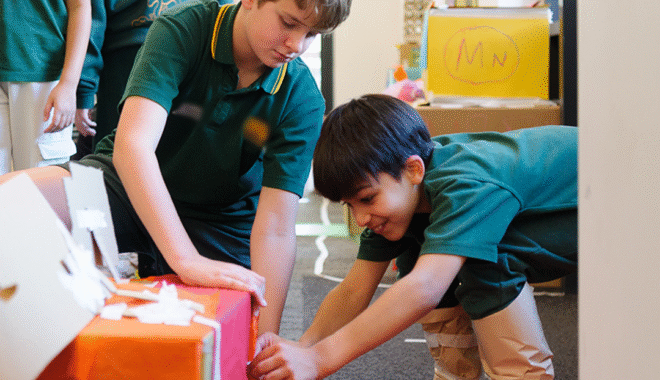 Wish Street production photo: Sarah Walker. Two children work together to attach paper to a box using tape. They both wear green school uniforms.