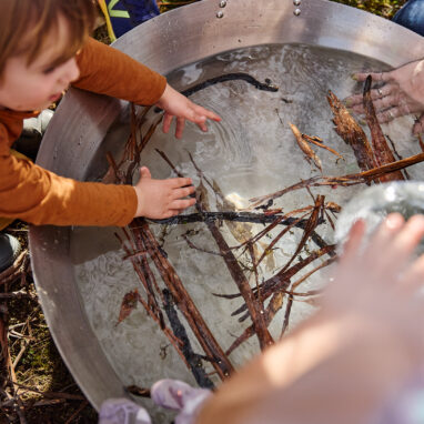 Forest production photo: Laura May Grogan. A young child in a brown t-shirt reaches into a large silver vat of water, filled with twigs, sticks and leaves.