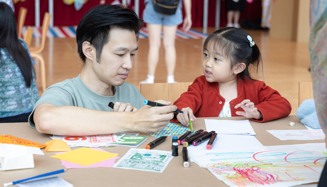 Pass It On production photo: Alvin Ho, Esplanade Singapore. A small child in a red cardigan and their adult create and play at a table with paper and coloured pastels. The child looks at the adult.