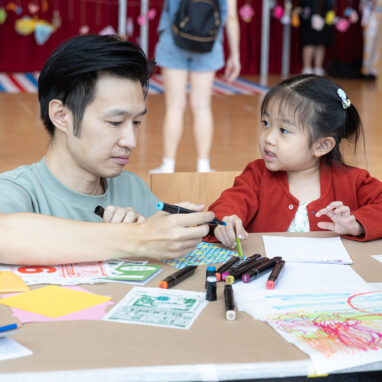 Pass It On production photo: Alvin Ho, Esplanade Singapore. A small child in a red cardigan and their adult create and play at a table with paper and coloured pastels. The child looks at the adult.
