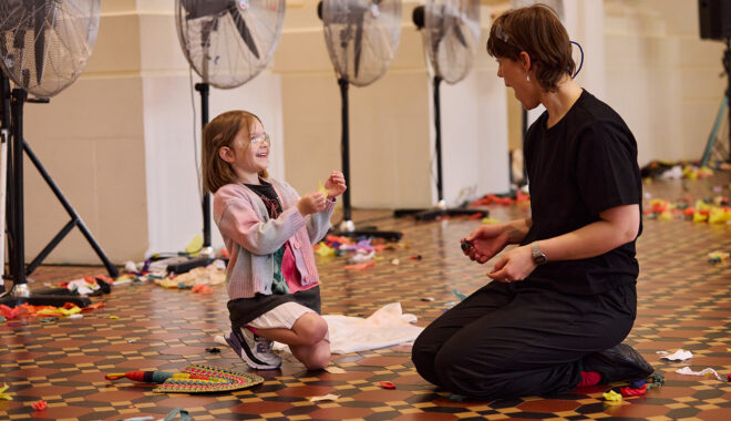 Whirlwind production photo: Laura May Grogan. A child wearing safety goggles and a Polyglot artist in black kneel on the floor together, creating and playing with paper. Large silver fans whir behind them.