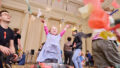 Whirlwind production photo: Laura May Grogan. A child in a lilac dress throws paper into the air over a silver fan. Their adult looks on.