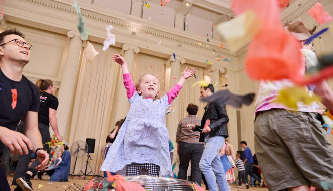 Whirlwind production photo: Laura May Grogan. A child in a lilac dress throws paper into the air over a silver fan. Their adult looks on.