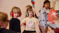 Whirlwind production photo: Laura May Grogan. Children wearing ear defenders smile at a Polyglot artist. Other children create and play behind them.
