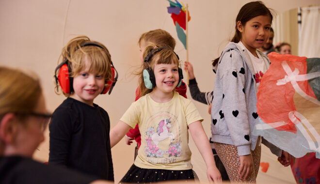 Whirlwind production photo: Laura May Grogan. Children wearing ear defenders smile at a Polyglot artist. Other children create and play behind them.