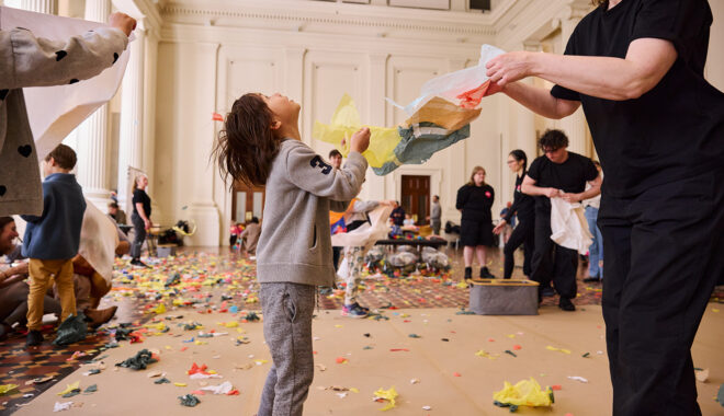 Whirlwind production photo: Laura May Grogan. A child in a grey outfit and a Polyglot artist in black create and play with pieces of tissue paper. The child looks up into the air. Other artists and families create and play in the background.