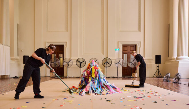 Whirlwind production photo: Laura May Grogan. An artist covered in strips of colourful tissue paper crouches in the centre of the performance space. Two artists wearing black sweep around them. There is a line of large silver fans in the background.