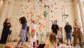 Whirlwind production photo: Laura May Grogan. Two children throw colourful paper confetti into the air. A third child lies on the floor watching. A Polyglot artist, an Auslan interpreter, and two adults with a baby also look on.