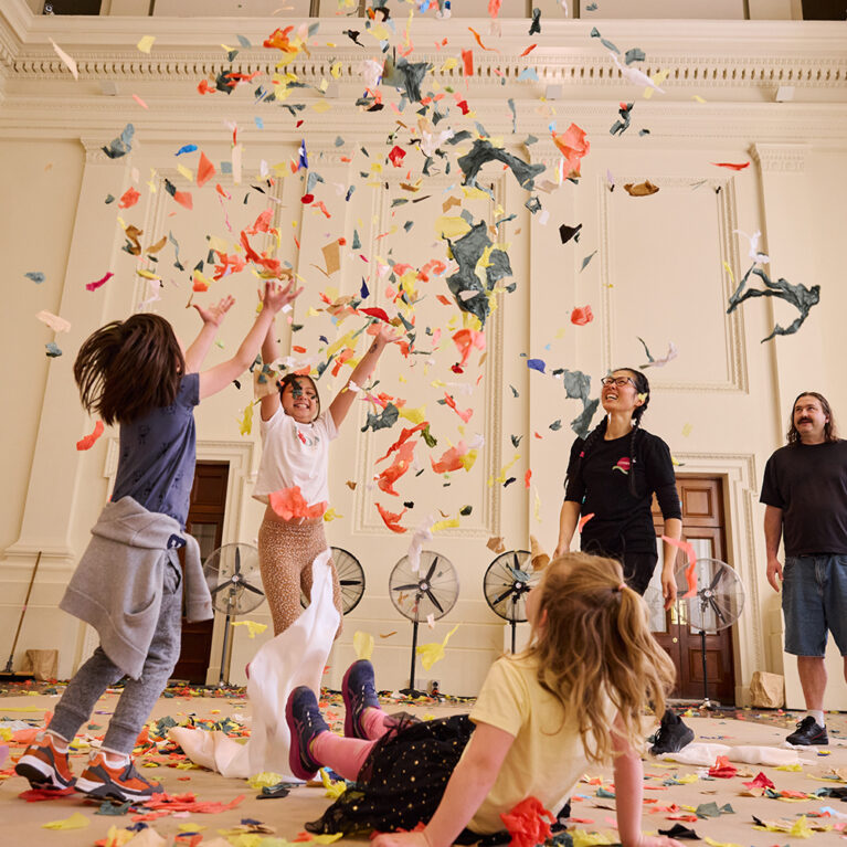 Whirlwind production photo: Laura May Grogan. Two children throw colourful paper confetti into the air. A third child lies on the floor watching. A Polyglot artist, an Auslan interpreter, and two adults with a baby also look on.