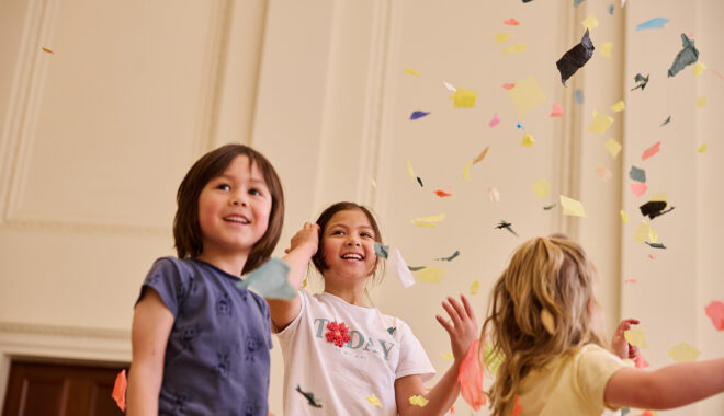 Whirlwind production photo: Laura May Grogan. Three children smile and play under a shower of colourful paper confetti.