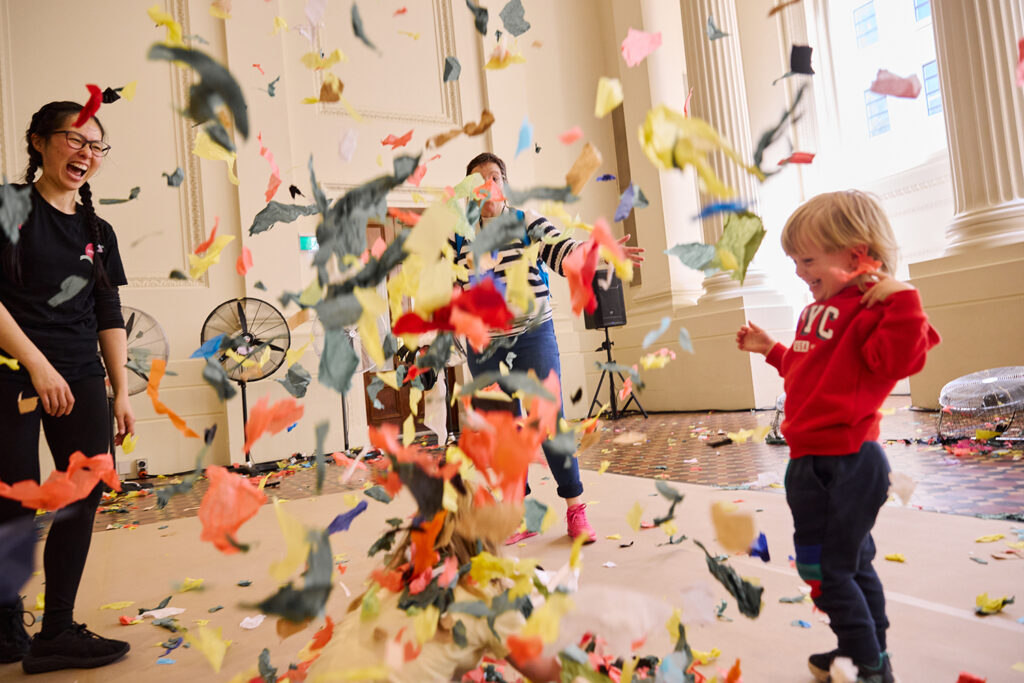 Whirlwind production photo: Laura May Grogan. A small child in a red jumper and a Polyglot artist laugh at a billowing cloud of colourful paper confetti.