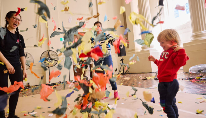 Whirlwind production photo: Laura May Grogan. A small child in a red jumper and a Polyglot artist laugh at a billowing cloud of colourful paper confetti.
