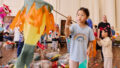 Whirlwind production photo: Laura May Grogan. A child in a blue t-shirt with a rainbow holds a large fish-shaped kite on a long wooden dowel. Children and families create and play in the background.