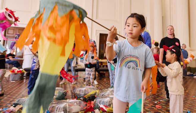 Whirlwind production photo: Laura May Grogan. A child in a blue t-shirt with a rainbow holds a large fish-shaped kite on a long wooden dowel. Children and families create and play in the background.