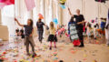 Whirlwind production photo: Laura May Grogan. Children and Polyglot artists move through the performance space holding kites and flags on long wooden dowels. The floor is covered in colourful paper scraps.