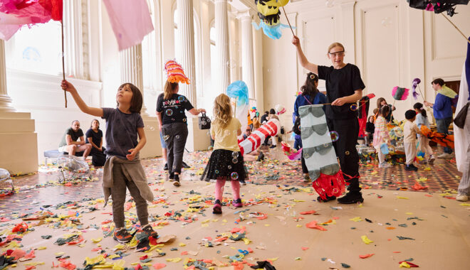 Whirlwind production photo: Laura May Grogan. Children and Polyglot artists move through the performance space holding kites and flags on long wooden dowels. The floor is covered in colourful paper scraps.