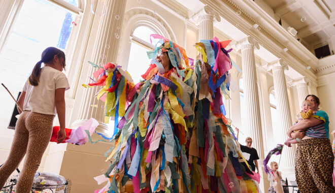 Whirlwind production photo: Laura May Grogan. A child and an adult holding a baby engage with a Polyglot artist who is covered in long strips of colourful tissue paper.