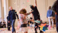 Whirlwind production photo: Laura May Grogan. A child holding a colourful pinwheel braces for a burst of colourful paper confetti. Polyglot artists and families create and play in the background.