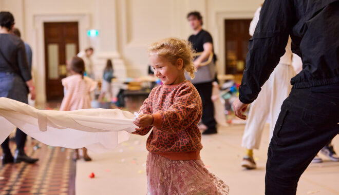 Whirlwind production photo: Laura May Grogan. A child holds a large white sheet. Polyglot artists and families create and play in the background.