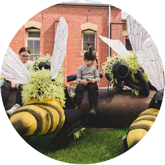 Bees production photo: Theresa Harrison. Polyglot artists in intricate Bee costumes are perched on and around a large metal pipe on green lawn. Children wearing handmade paper antennae and wings sit on the pipe amongst them. There is a heritage red brick building in the background.