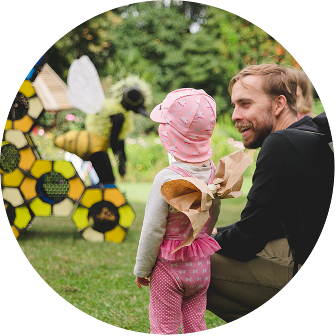 Bees production photo: Theresa Harrison. A child wearing pink and paper wings looks toward a Bee. Their adult is crouched beside them,