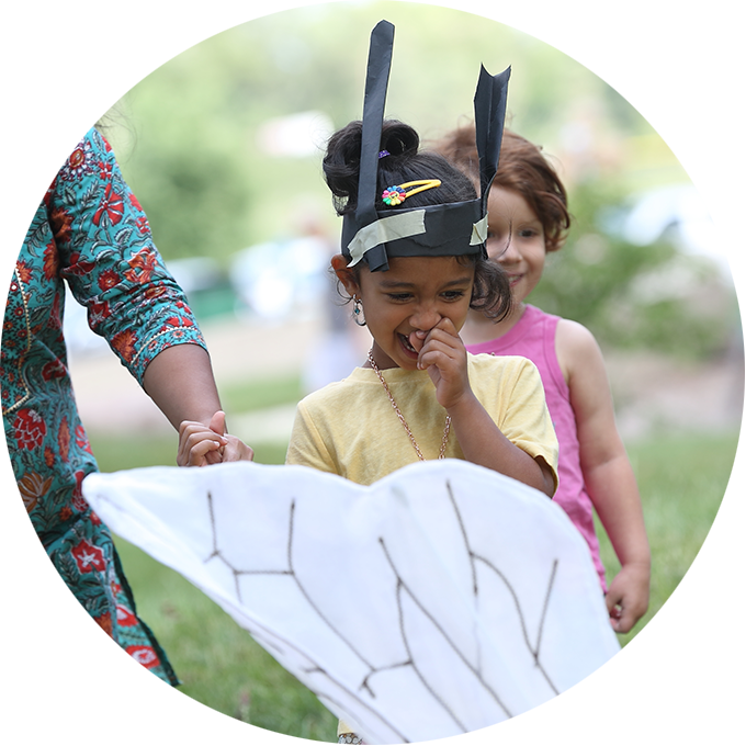 Bees production photo. Dan Welk. Two children, one wearing handmade paper antennae and holding their adult’s hand, joyfully engage with an artist in a Bee costume (only part of their wing is visible).