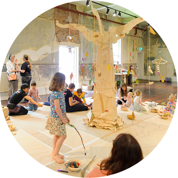 Art Tree production photo: Sarah Walker. A child wearing a blue shirt is holding a stick, and standing in the centre of the frame. There's a large cardboard tree in the background with families and artists sitting or standing around it.