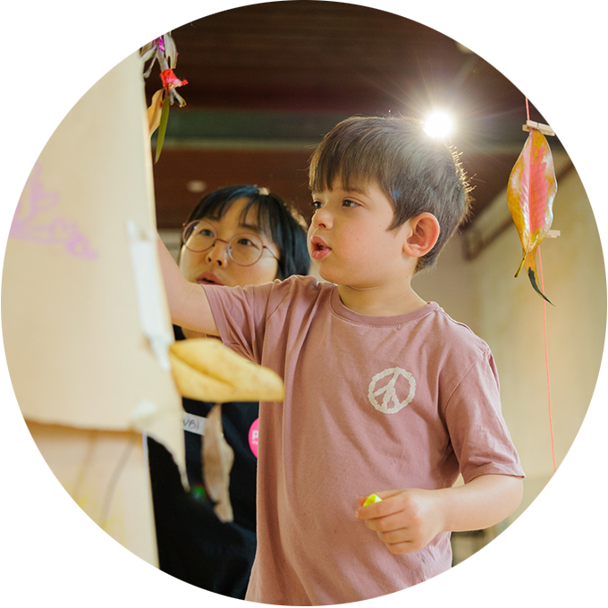 Art Tree production photo. A child and a Polyglot artist use markers to add to a brown paper tree. Colourful, decorated leaves and twigs are suspended with yarn. The child’s adult looks on. Photo: Sarah Walker