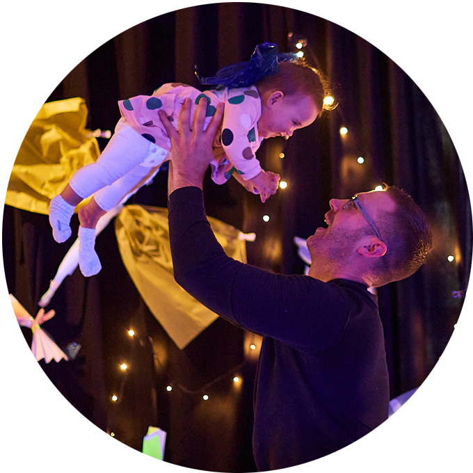 Catch the Light production photo: An adult lifts a young child in the air. There is a purple lighting wash on them, and lights in the background. Photo: Laura May Grogan