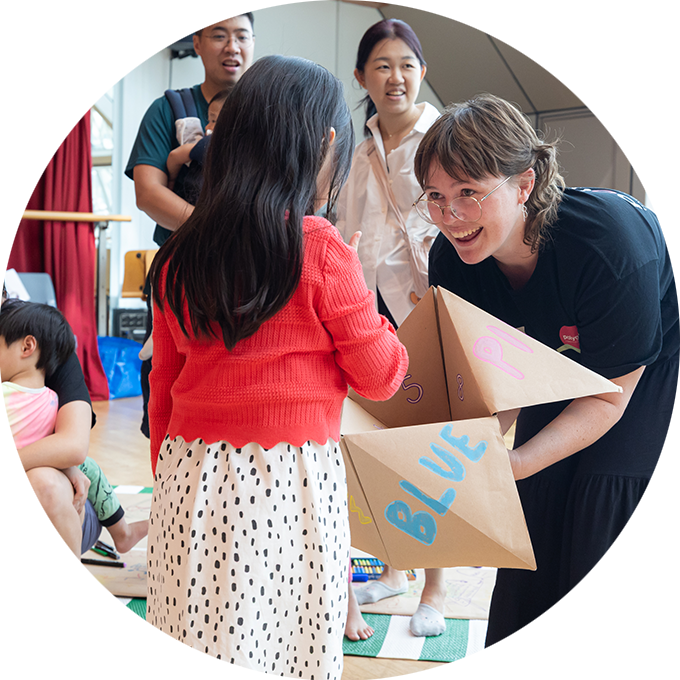 Pass It On workshop photo: Alvin Ho. A Polyglot artist leans forward holding a large paper chatter box. They are facing a child in an orange sweater. There are two adults in the background.