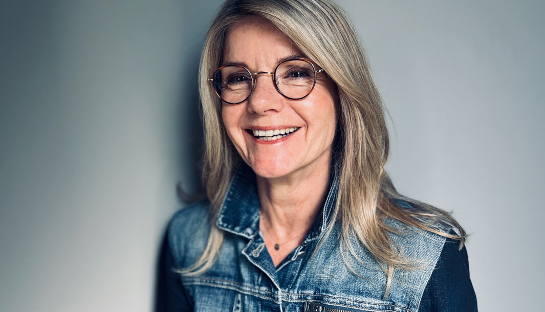 A portrait of Ally Harvey. She has blonde hair, and wears round glasses and a denim jacket. She is in front of a grey background, smiling off-camera.