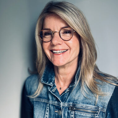 A portrait of Ally Harvey. She has blonde hair, and wears round glasses and a denim jacket. She is in front of a grey background, smiling off-camera.