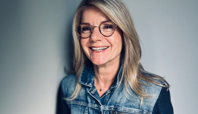 A portrait of Ally Harvey. She has blonde hair, and wears round glasses and a denim jacket. She is in front of a grey background, smiling off-camera.