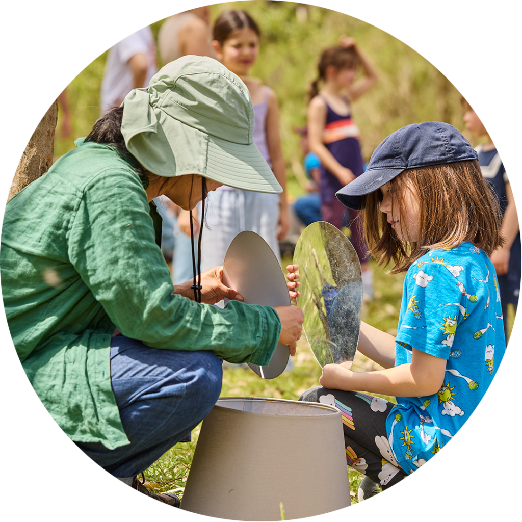Forest production photo: Laura May Grogan. A child in a blue t-shirt sits across an adult in a green blouse. They inspect a handheld round mirror together. There are children and families in the background.
