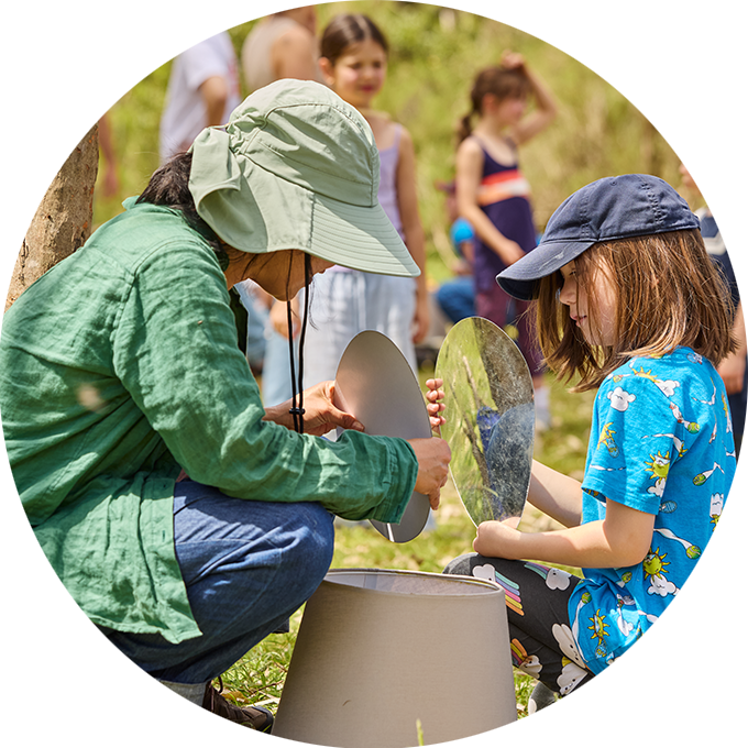 Forest workshop photo: A Polyglot artist plays with a child, both holding mirrors in the forest. There are children in the background. Photo: Laura May Grogan.