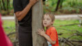 Forest production photo: Claudia Sangiorgi Dalimore. A child in an orange t-shirt presses their ear to a tree trunk, listening. A Polyglot artist wearing black stands on the other side of the tree, holding the trunk.