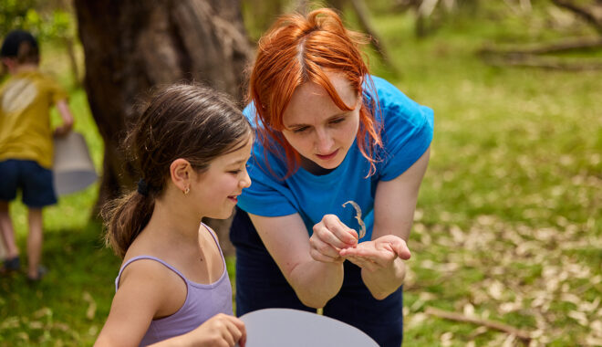 Forest production photo: Laura May Grogan. A Polyglot artist and a child look closely at a leaf. They are in a bushland clearing.