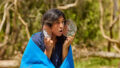 Forest production photo: Laura May Grogan. A Polyglot artist draped in a large blue blanket holds small round mirrors to their face, similar to fish gills/fins. Their mouth is open. They are in a bushland clearing.