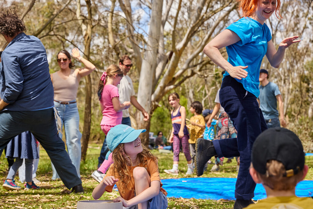 Forest production photo: Laura May Grogan. Polyglot artists dance in a bushland clearing. Children and families around them watch, or join in.